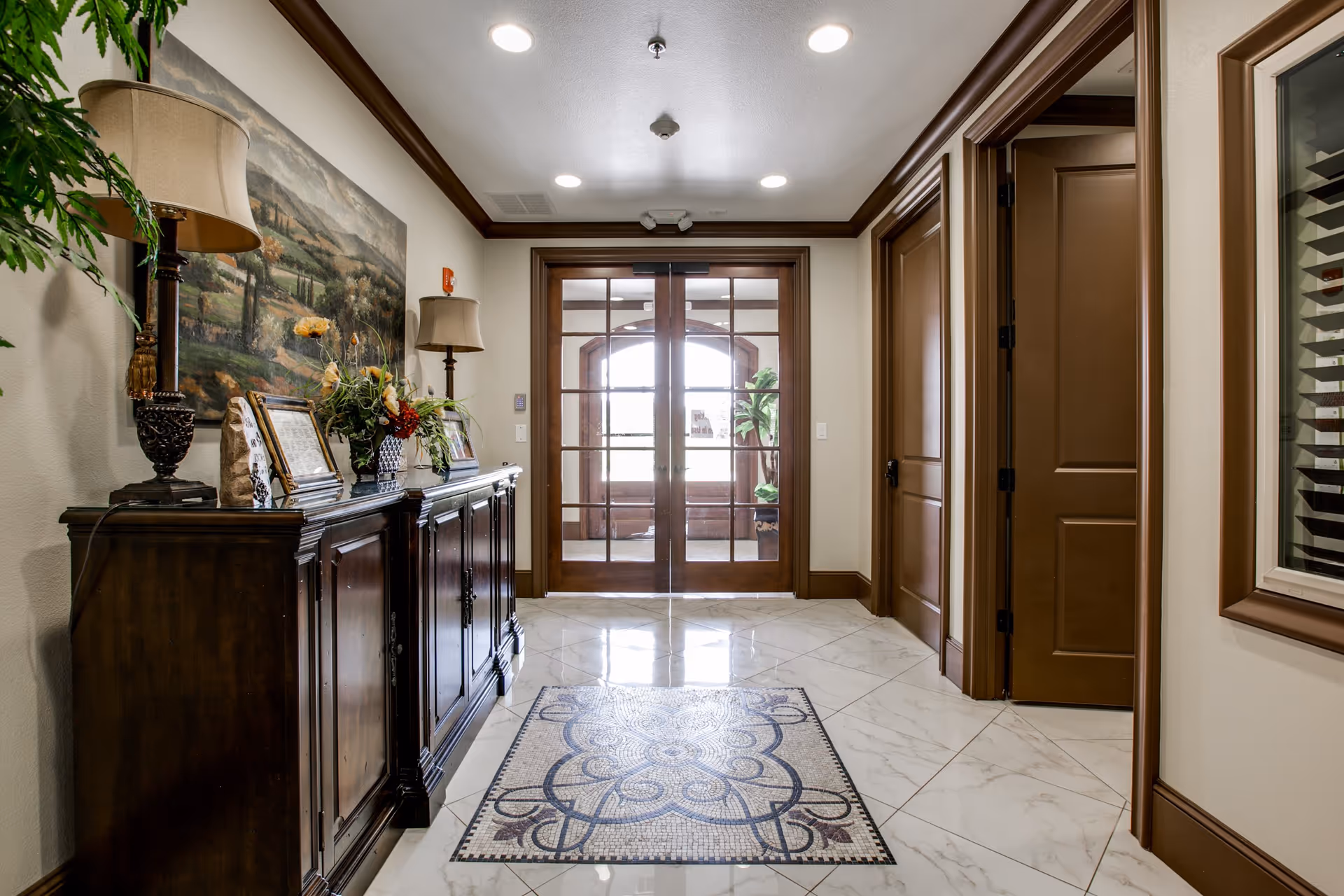 A well-lit hallway with polished white marble floors featuring a decorative tile pattern in the center. On the left side, there is a dark wooden cabinet adorned with two table lamps, framed pictures, and a floral arrangement. Above the cabinet hangs a large landscape painting. The hallway ends with double glass doors framed in dark wood, leading to another room with a large window and plants. On the right side, there are two closed brown wooden doors and a window with wooden blinds.