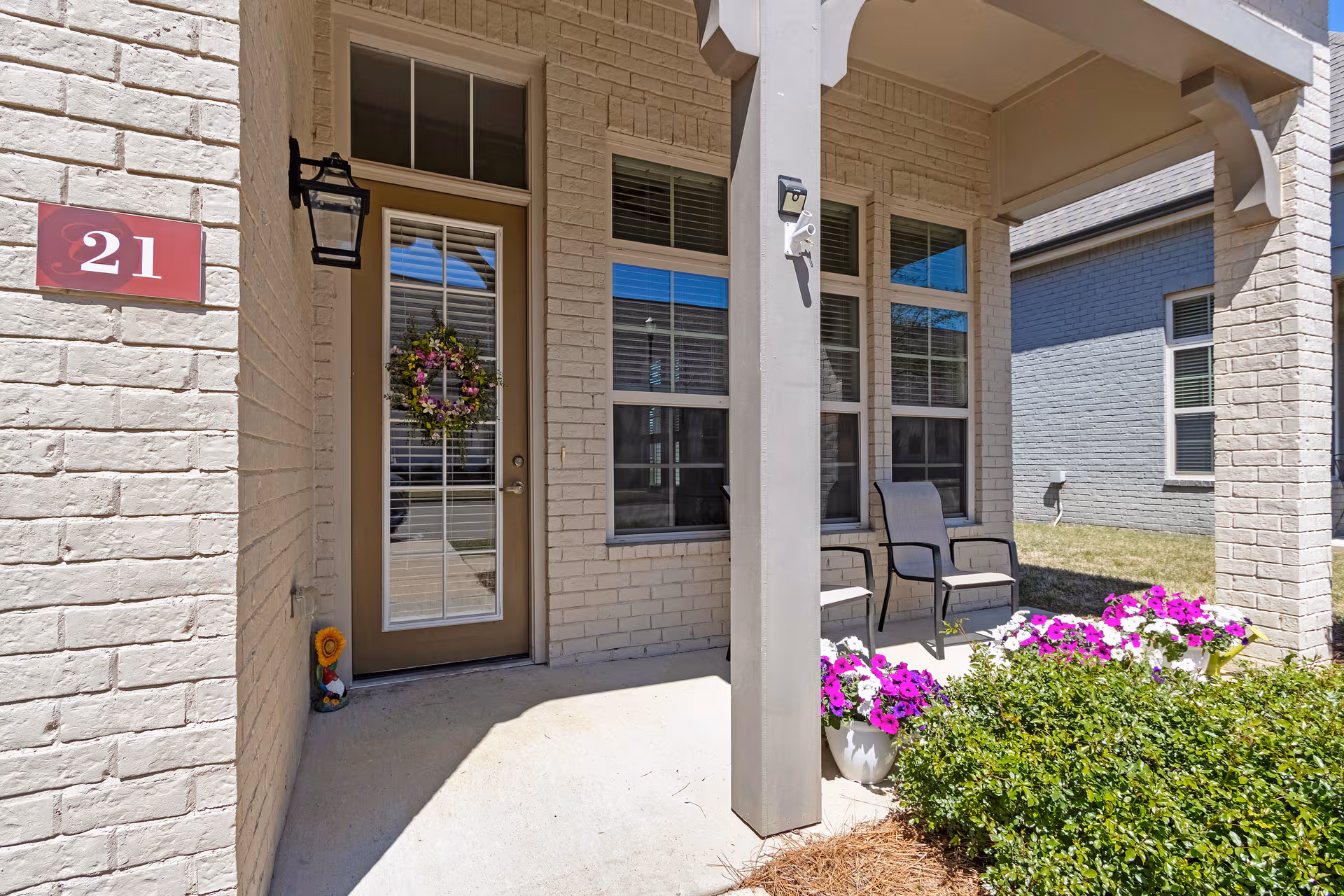 Brick building entrance with door, wreath, porch chairs, and vibrant flowers.