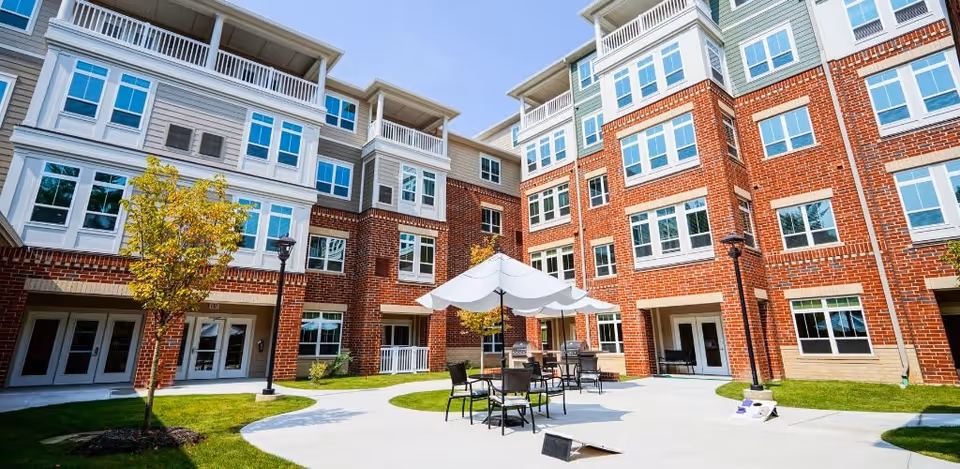 Outdoor courtyard area of a senior living facility with brick and white-paneled buildings surrounding a paved patio. The patio has tables with umbrellas and chairs, small trees, and a cornhole game set up on the ground under a clear blue sky.