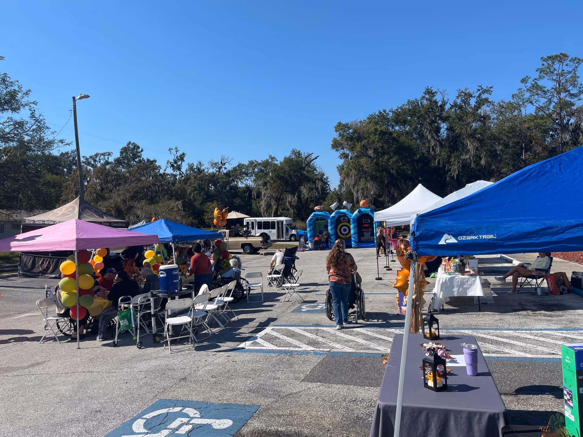 Outdoor community fair with tents, balloons, tables, and people gathered in a parking lot.