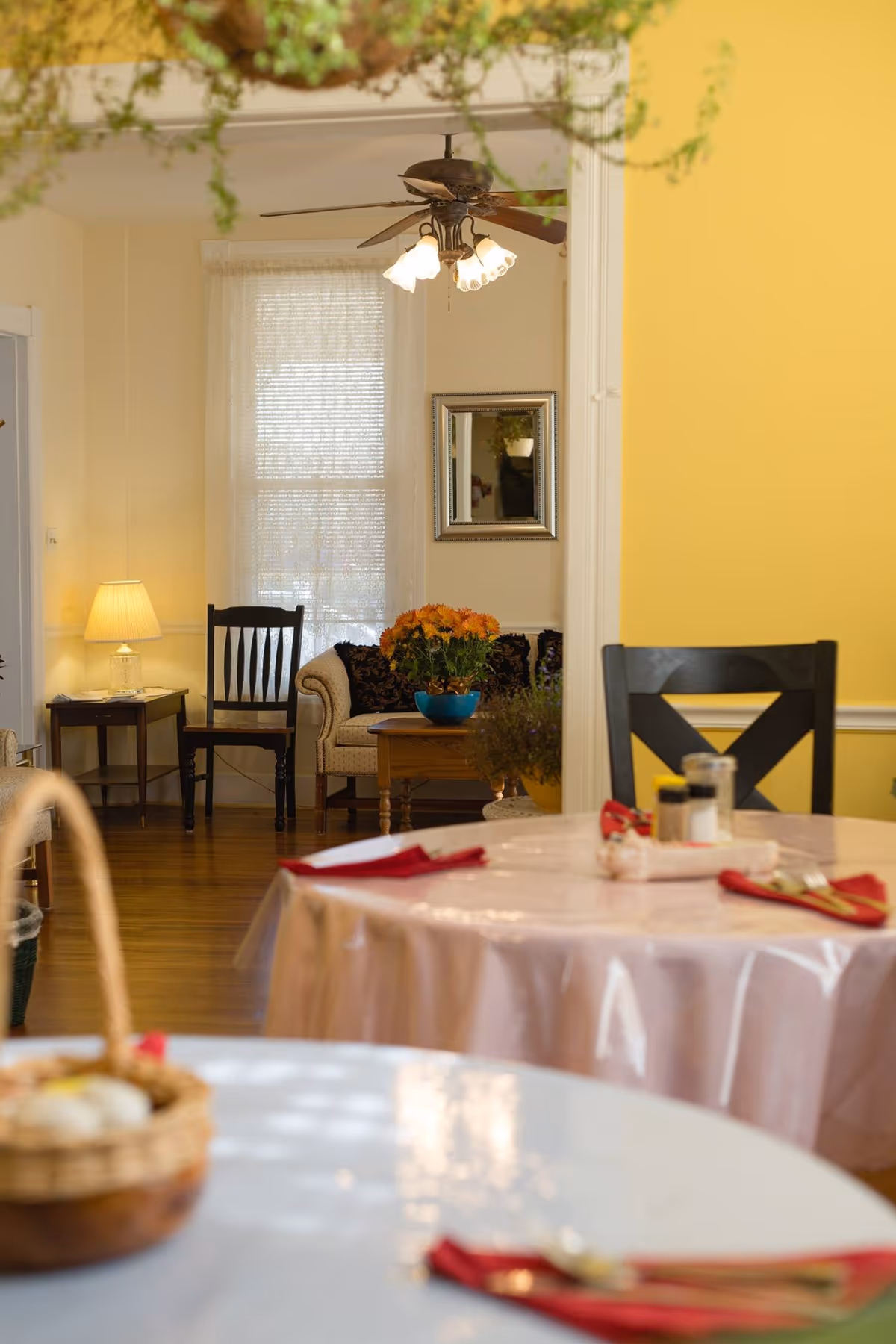 Interior view of a senior living facility dining area with round tables covered in plastic tablecloths, set with red napkins and silverware. In the background, there is a cozy sitting area with a beige couch, a wooden chair, a side table with a lamp, a ceiling fan with lights, and a window with sheer curtains. A vase with orange flowers is on the coffee table.