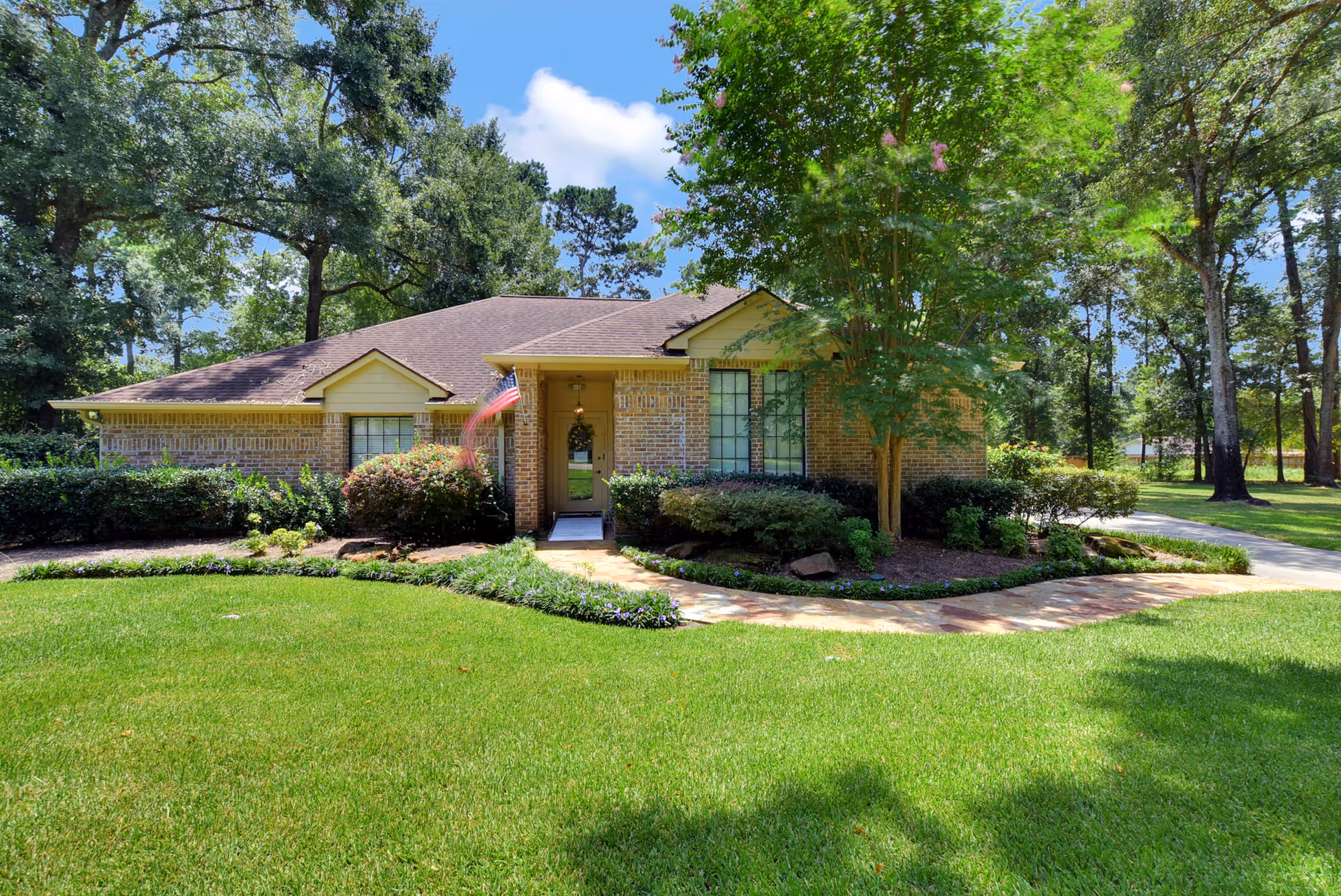 Front exterior view of a single-story brick house with a well-maintained lawn, a stone pathway leading to the front door, bushes and trees surrounding the house, and an American flag near the entrance under a clear blue sky.