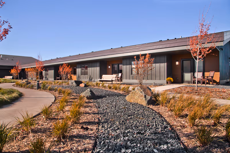 Exterior view of a senior living facility with a winding path, rocks, and outdoor seating.