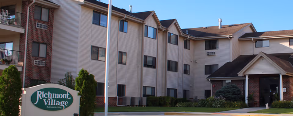 Exterior view of Richmont Village retirement living facility showing a multi-story building with beige siding and brick accents, multiple windows, and a main entrance with a small covered porch. There is a green and white sign in front with the facility name and some landscaping including bushes and trees.