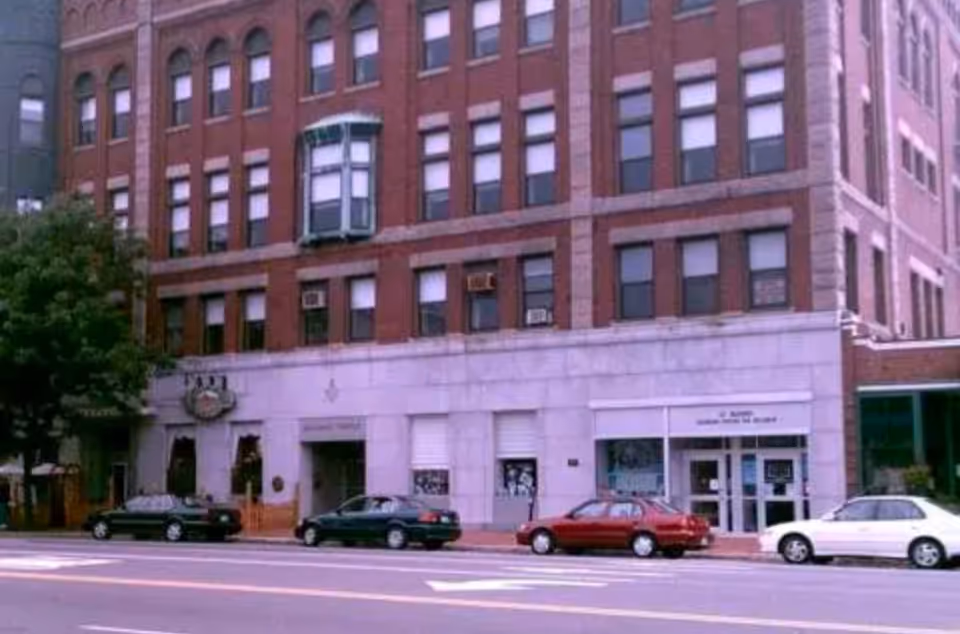 Brick, multi-story building with storefronts on a city street and parked cars.