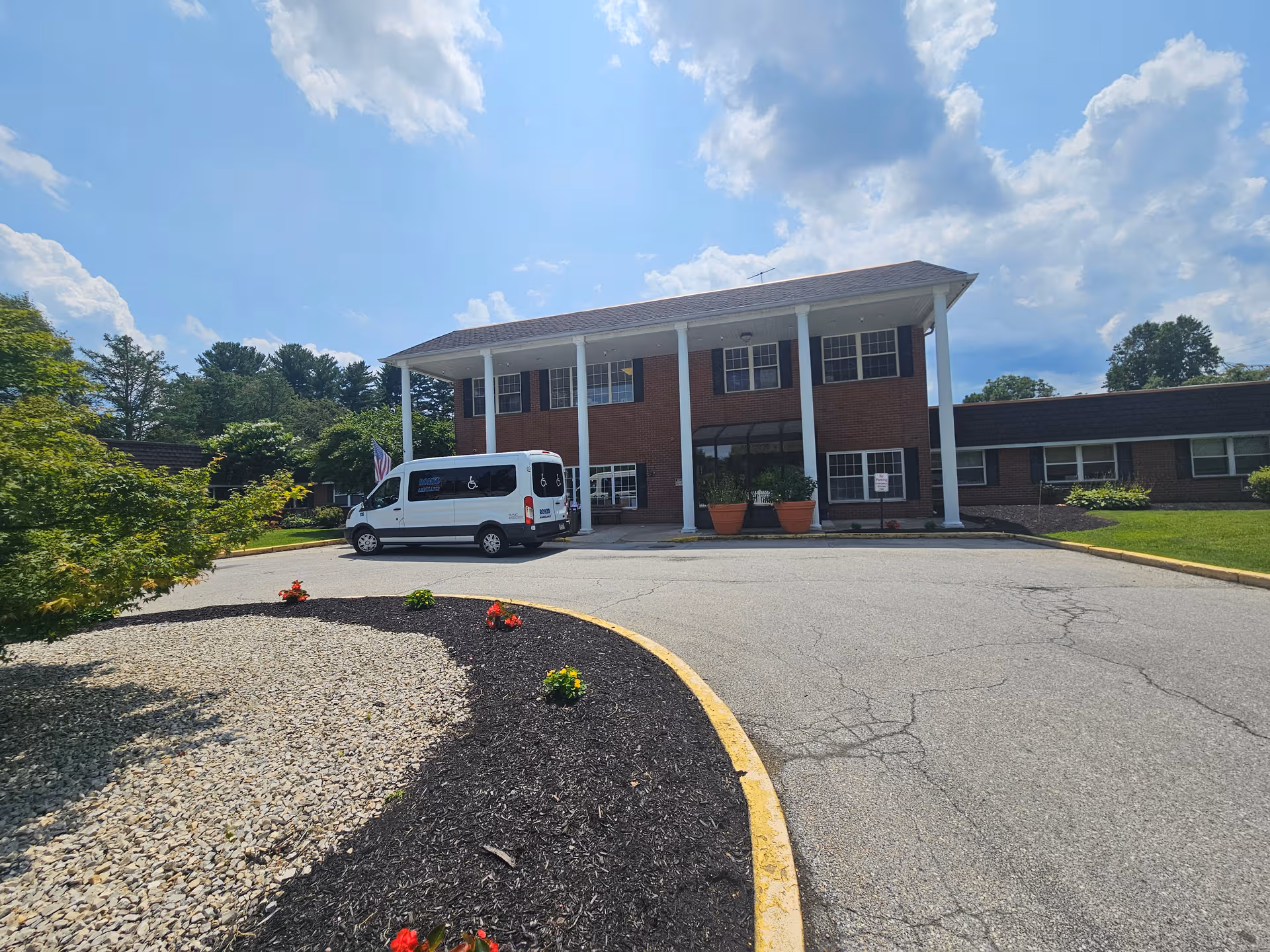 Brick senior living facility with white columns, van parked at entrance, landscaped circular driveway.