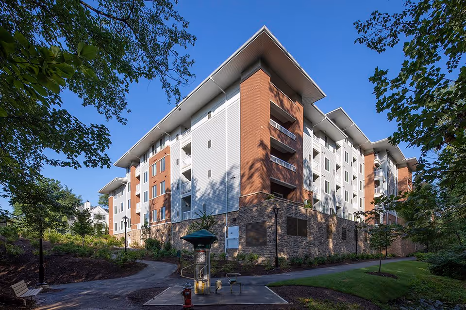 Exterior view of a multi-story senior living facility building with brick and white siding, surrounded by trees and greenery under a clear blue sky. There is a paved path, a bench, and a fire hydrant in the foreground.