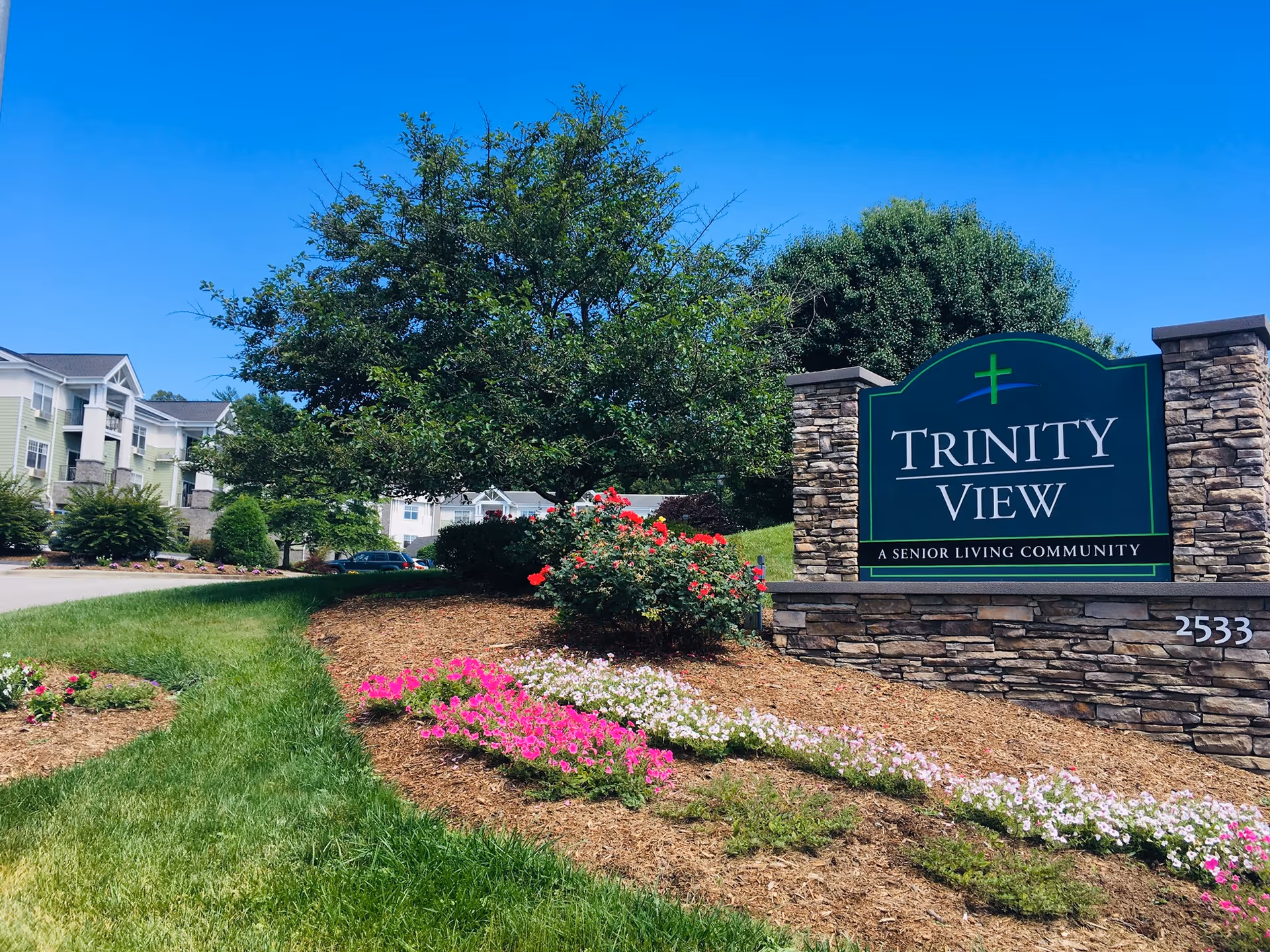 Entrance sign for Trinity View senior living community with landscaped flower beds, trees, and apartment buildings in the background.