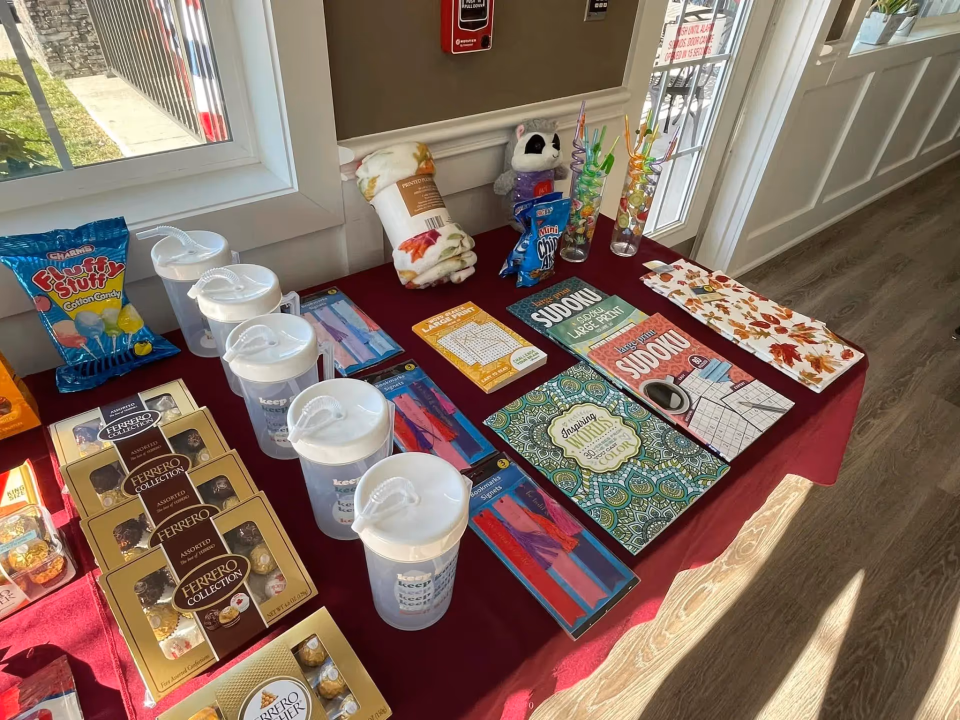 Indoor hallway table with brochures, chocolates, cups, candy, games, and a plush toy near windows.
