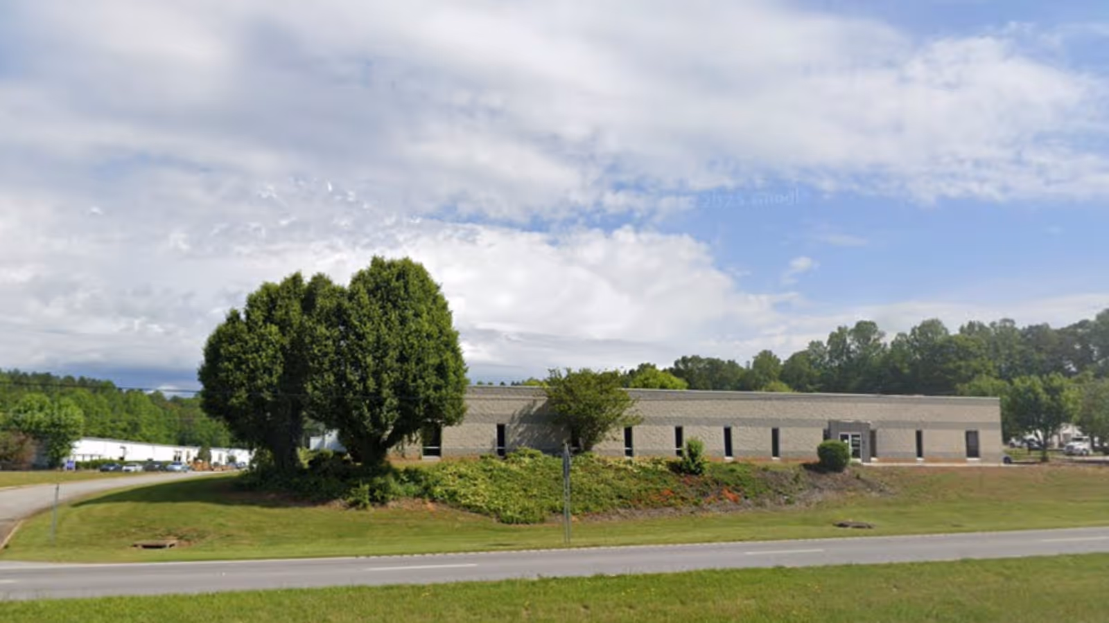 Exterior view of a single-story building with trimmed lawn, trees, and a road in front.