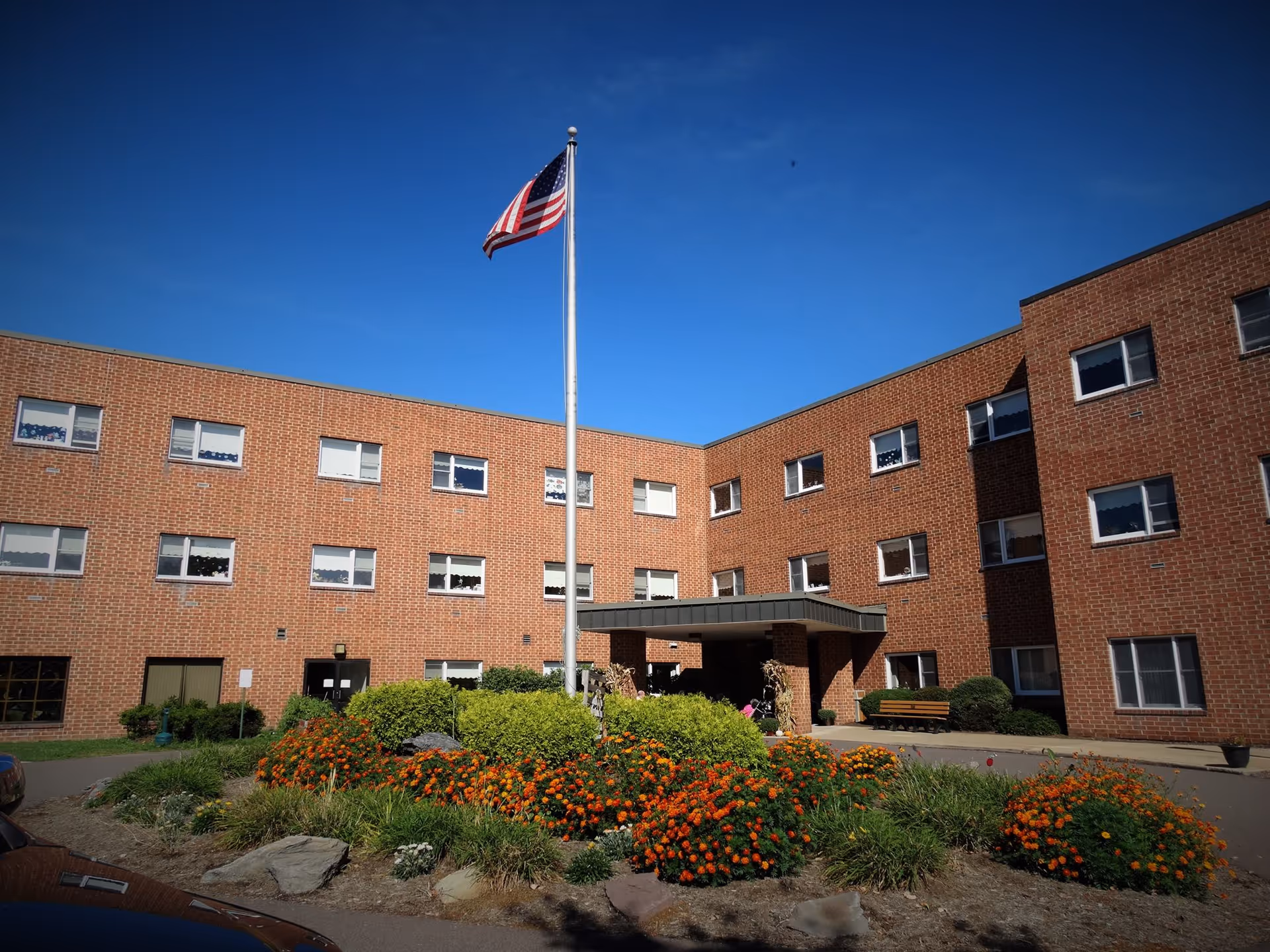 Brick nursing home exterior with flagpole, flowering landscaping, and a covered entrance.