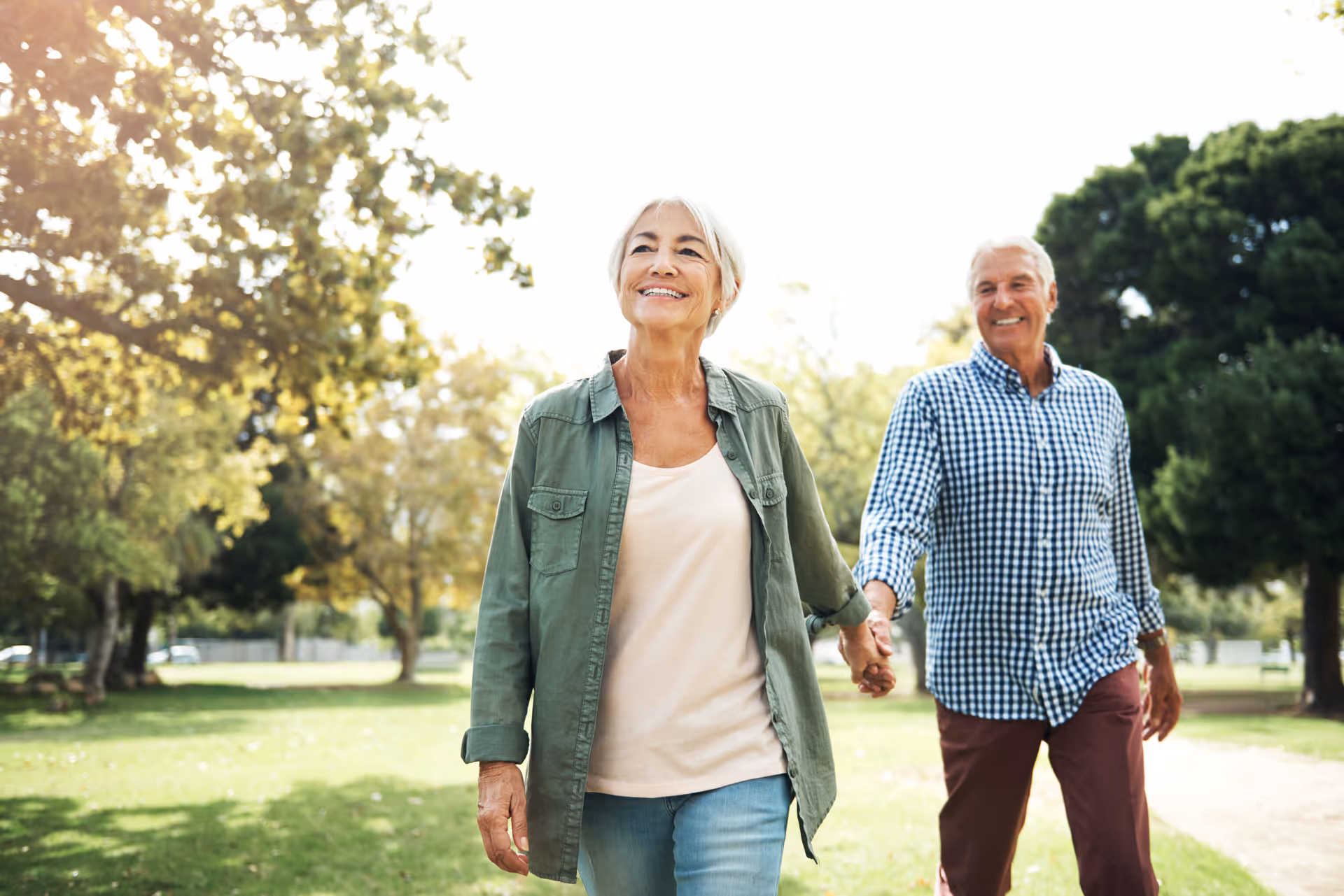 An elderly couple holding hands and walking outdoors in a park-like setting with trees and grass, smiling and enjoying a sunny day.