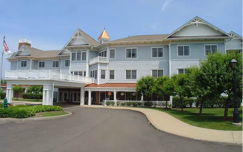Exterior view of a large, multi-story senior living facility building with white siding, multiple windows, a covered entrance, and a driveway surrounded by green trees and landscaping under a clear blue sky.