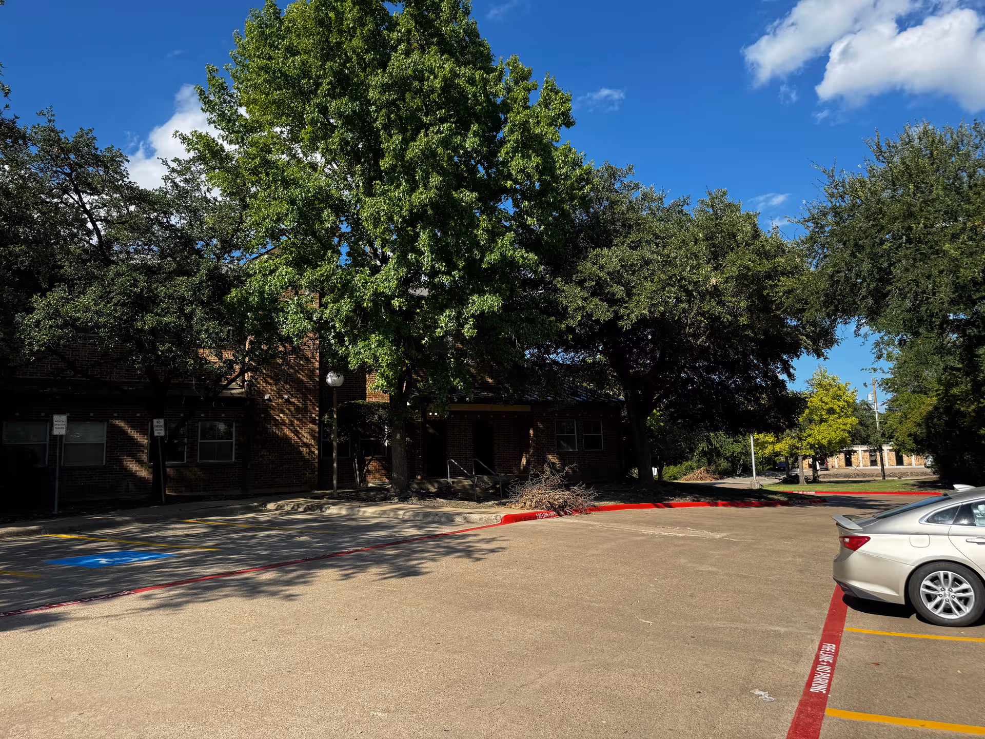 Brick senior living facility partially obscured by large trees, with a parking lot in foreground.