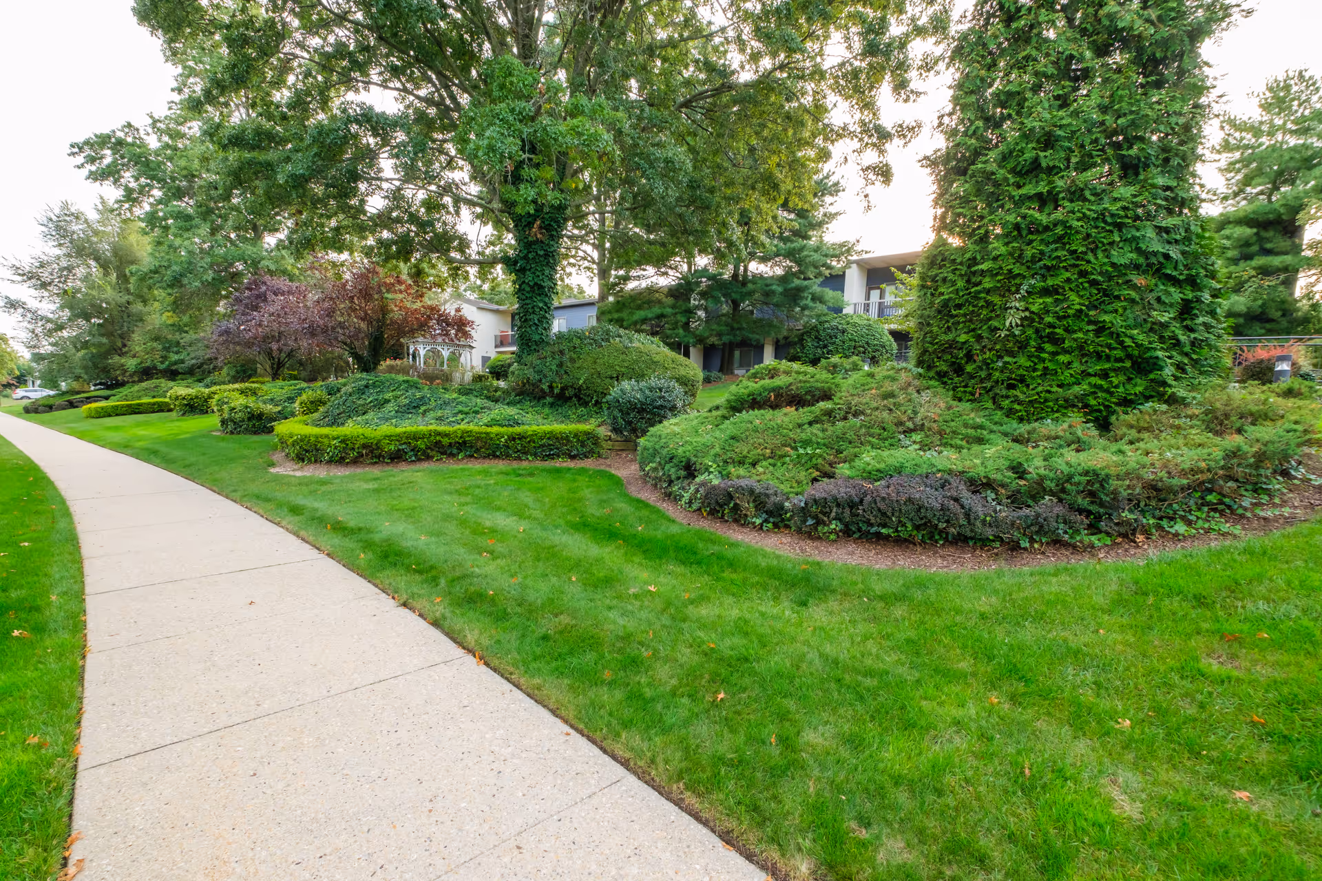 A curved sidewalk passes manicured lawns, shrubs and trees in front of a low-rise residential building.