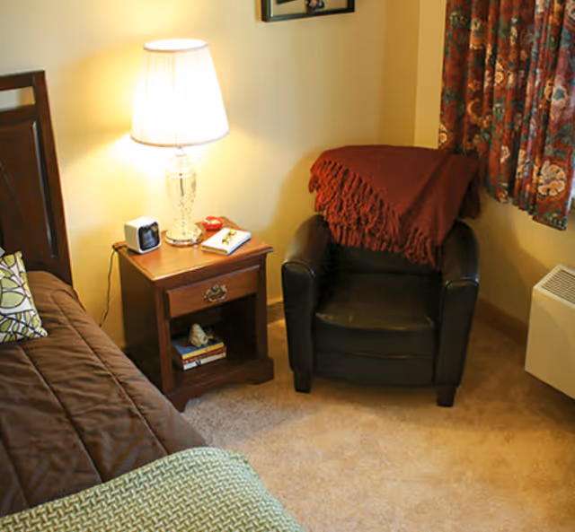 Cozy bedroom corner with a bed, wooden nightstand and lamp, and a black armchair draped with a red throw.