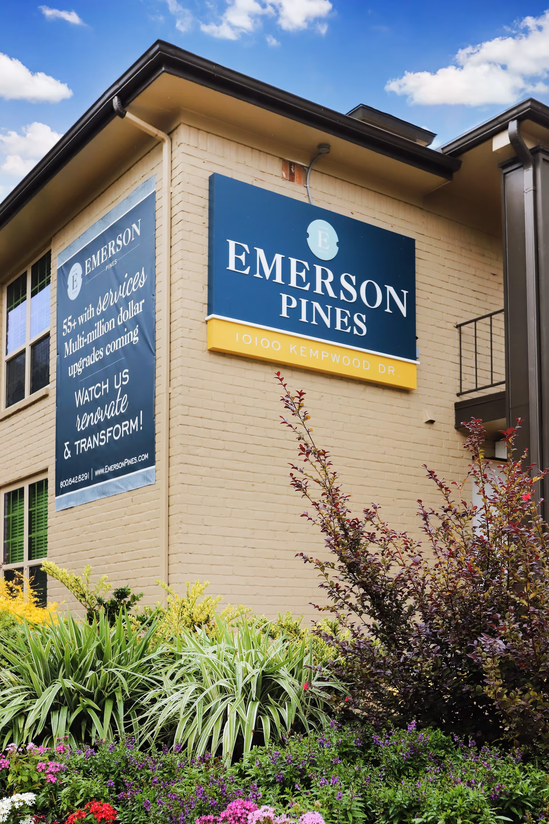 Exterior facade of a building with a large blue "Emerson Pines" sign and landscaped flowers in front.