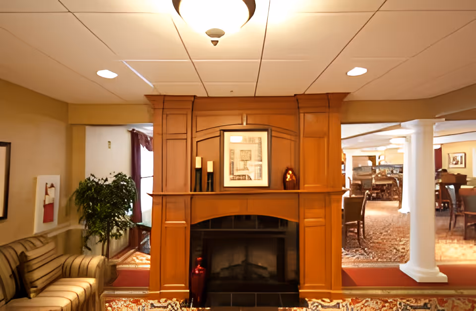 Cozy interior of a senior living facility featuring a wooden fireplace with decorative items on the mantel, a striped sofa on the left, a potted plant, and a view into a dining area with tables and chairs supported by white columns.
