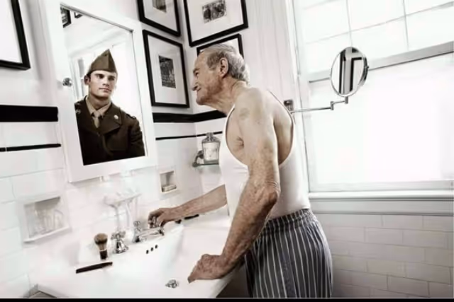 Elderly man at a bathroom sink gazes at a wall of framed photos.