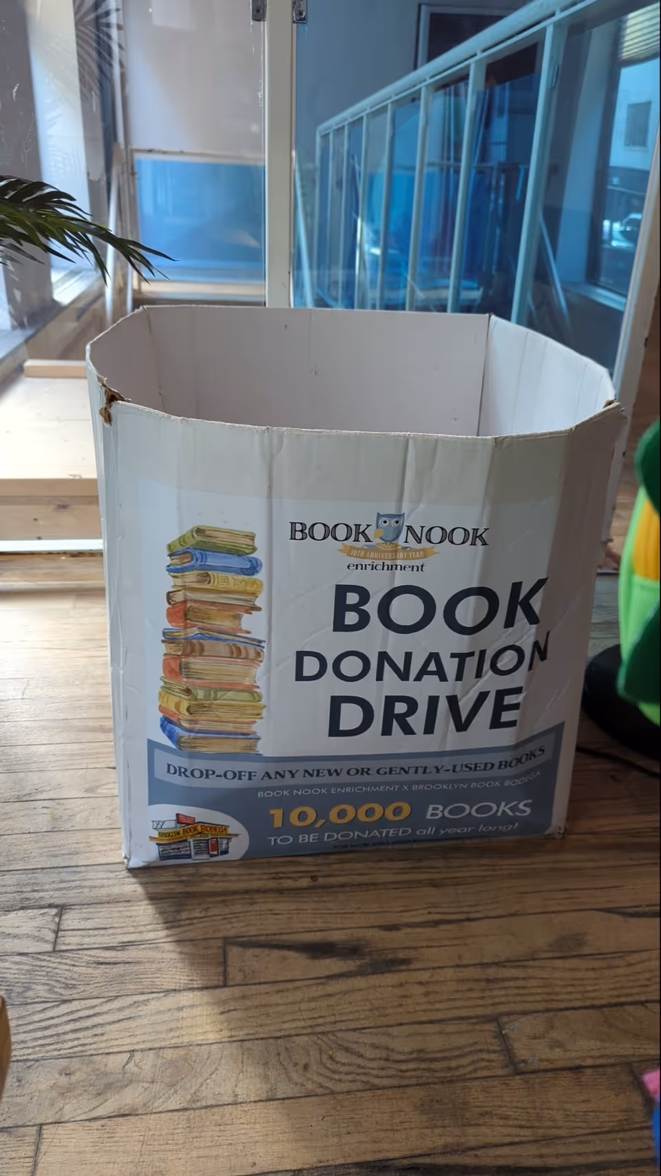Cardboard book donation box on a wooden floor inside a facility lobby