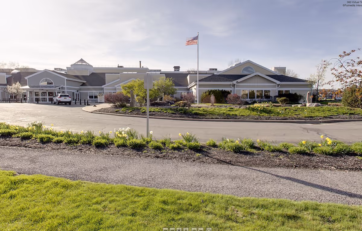 Exterior view of Dosberg Manor Adlt Hme-Menorah building with a circular driveway, landscaped garden with flowers and shrubs, an American flag on a flagpole, and a clear sky.