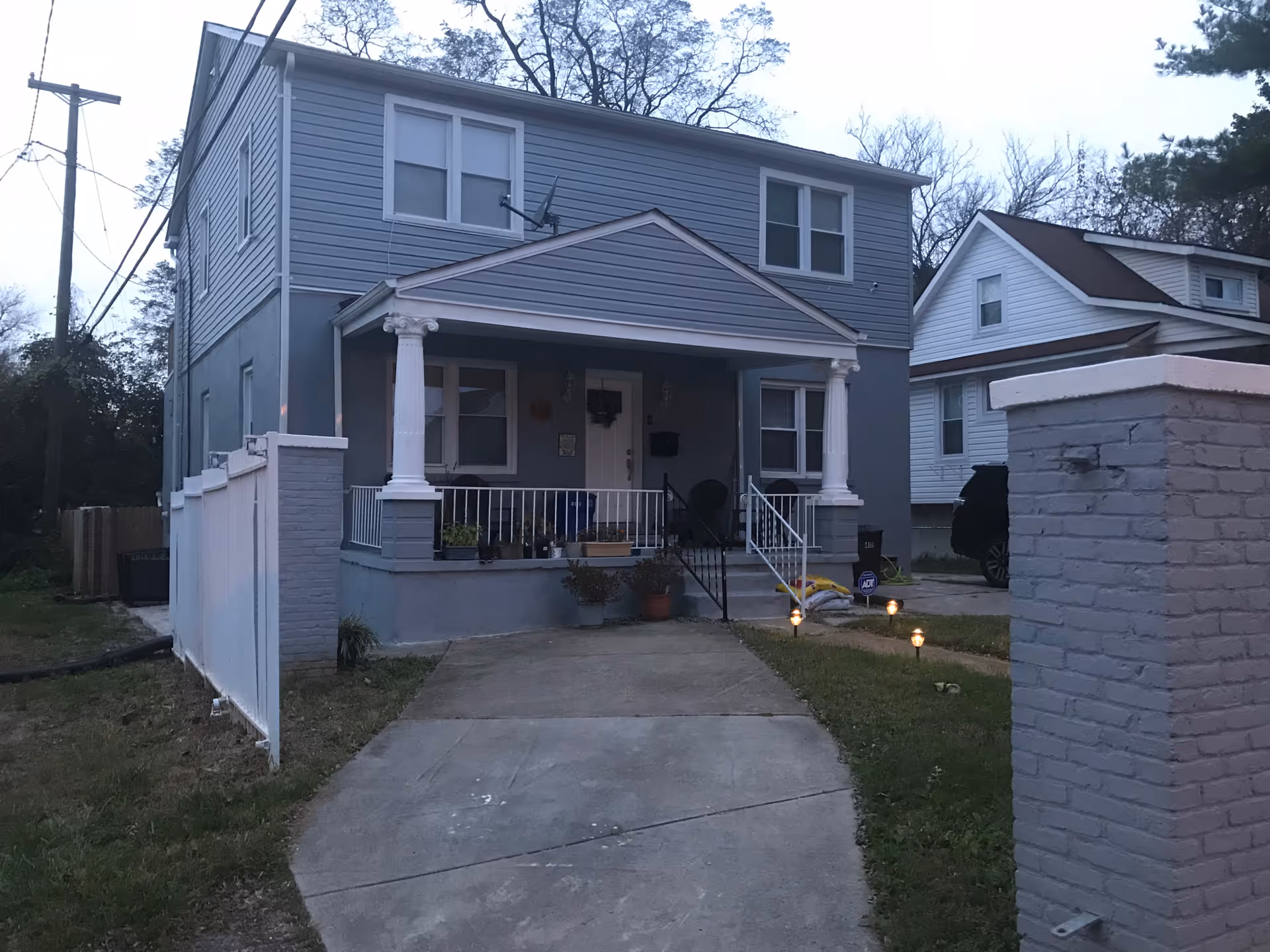Front exterior view of a two-story gray house with white trim and a covered porch supported by two white columns. There is a concrete driveway leading up to the porch, a white fence on the left side, and a small lawn with pathway lights on the right. Another house is visible in the background on the right.