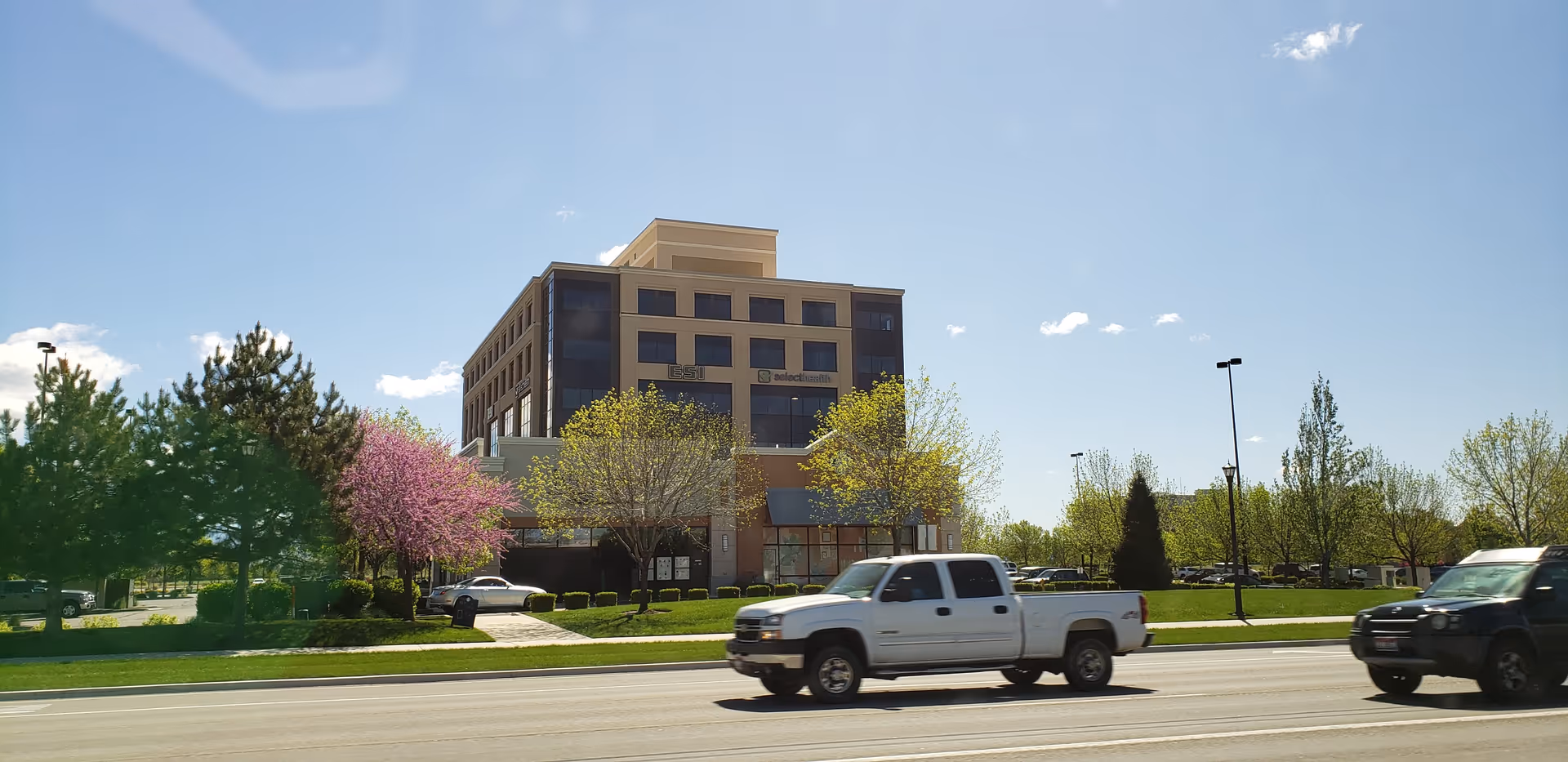 Select Health senior living building exterior seen with trees, parked cars, and a clear blue sky.