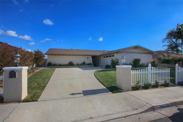 A single-story senior living facility with a long driveway, lawn, and white picket fence.