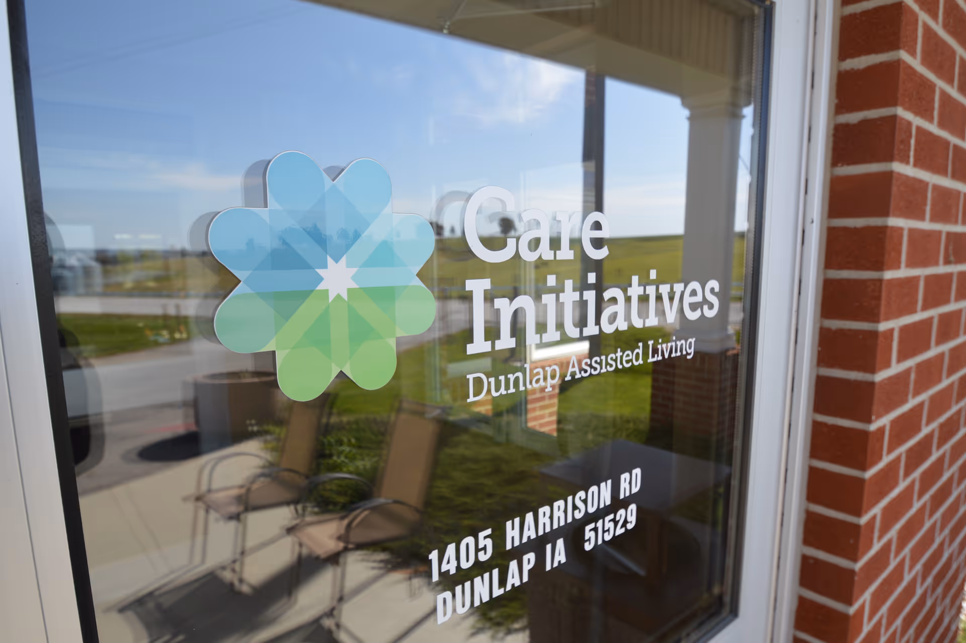 Glass entrance door with the 'Care Initiatives Dunlap Assisted Living' logo and address, reflecting outdoor chairs and lawn.
