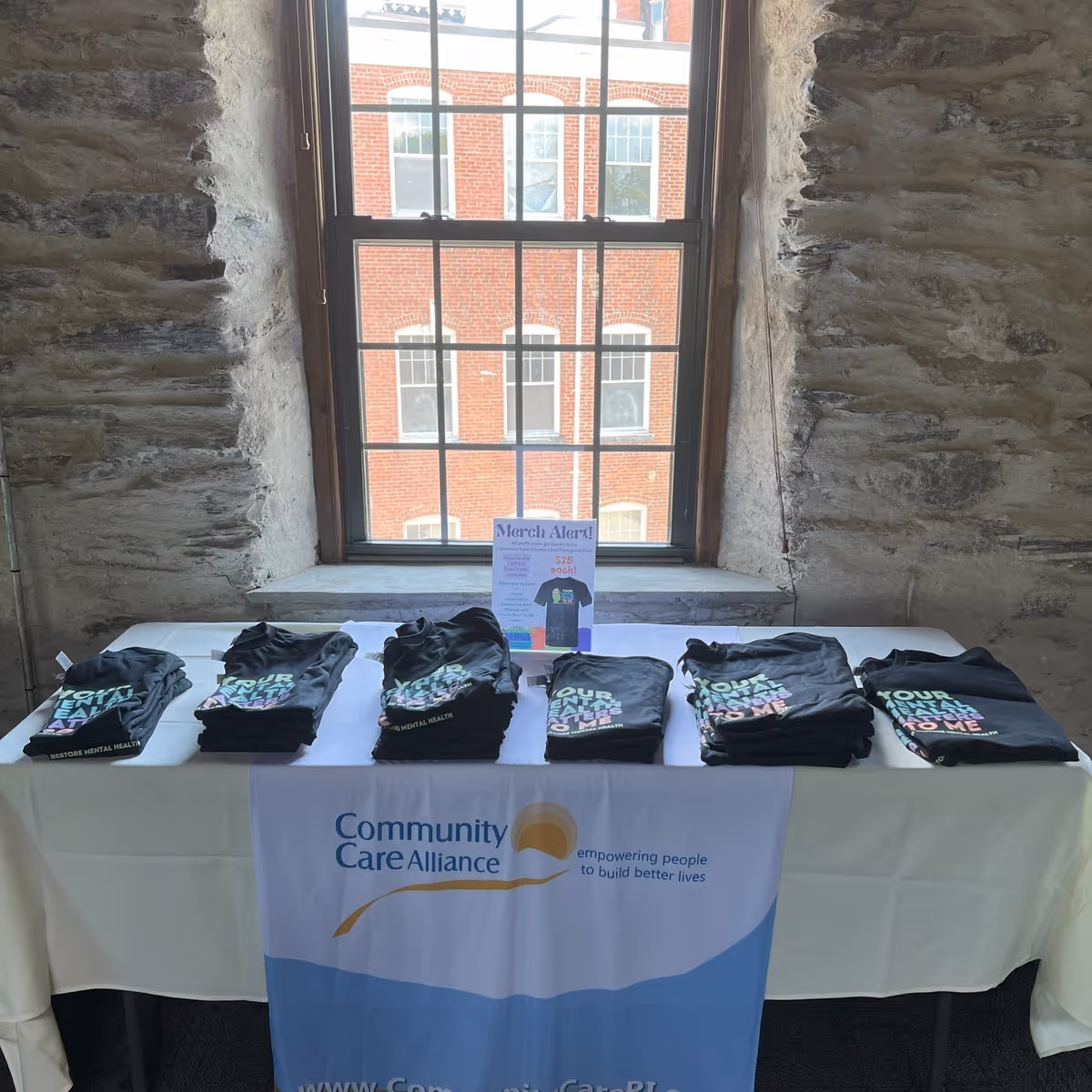 Table displaying folded t-shirts and a Community Care Alliance banner in front of a window set in a stone-walled interior.