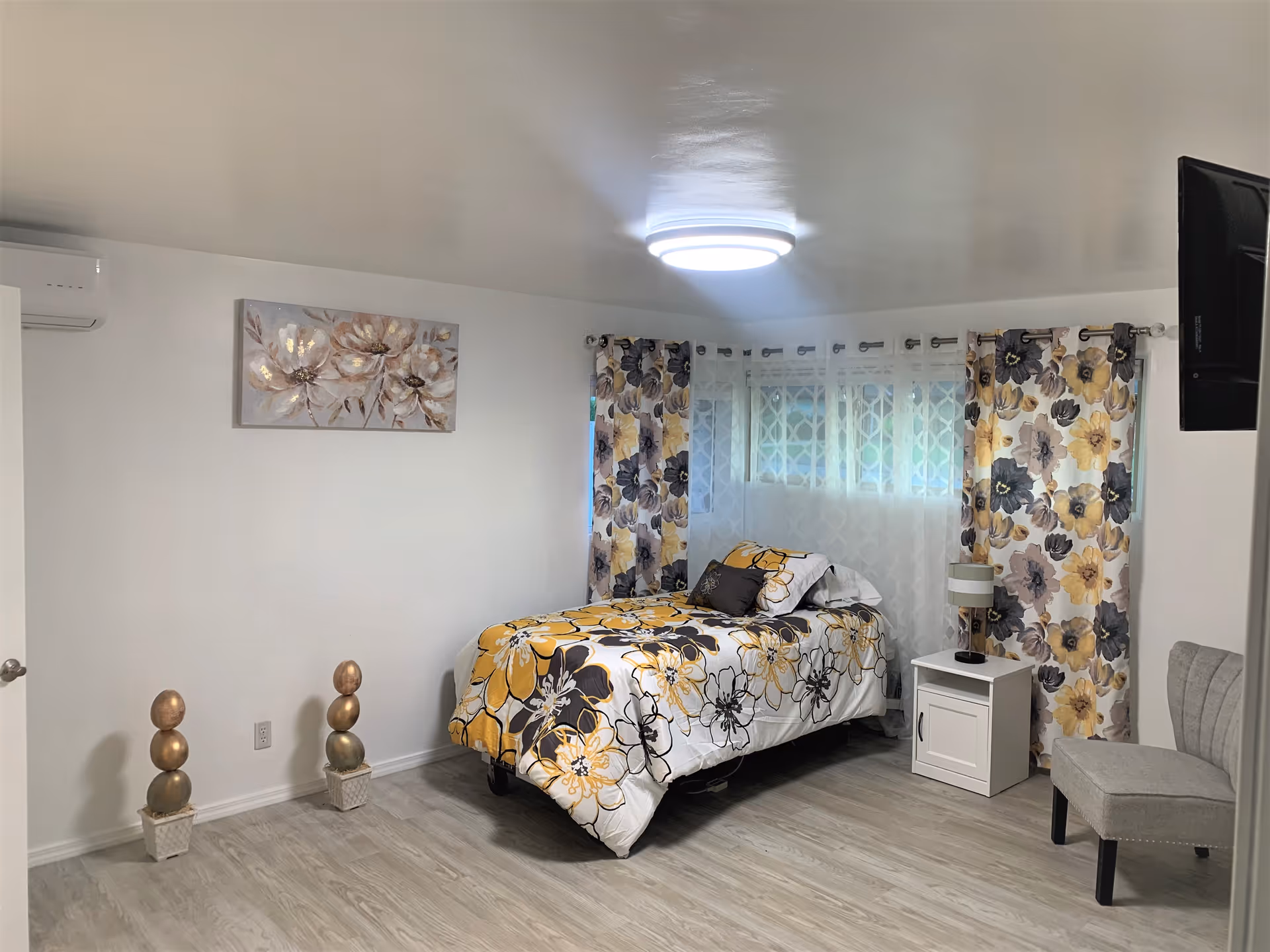 Bright bedroom with floral bedding, patterned curtains, nightstand, and a chair against white walls.