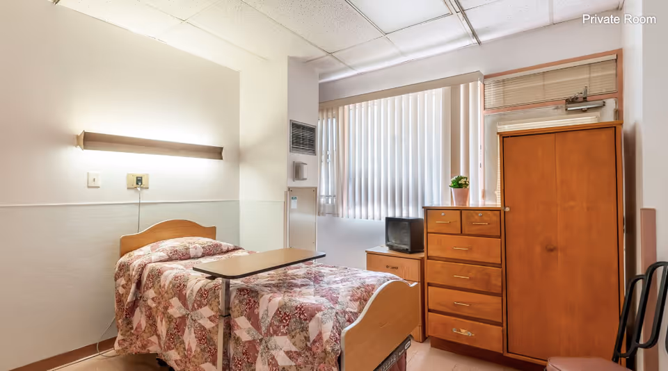 A furnished private bedroom in a senior facility with a single bed, overbed table, dresser, wardrobe, and a window with vertical blinds.