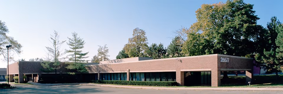 Single-story brick senior living facility exterior with large windows, trees, and a surrounding parking area.
