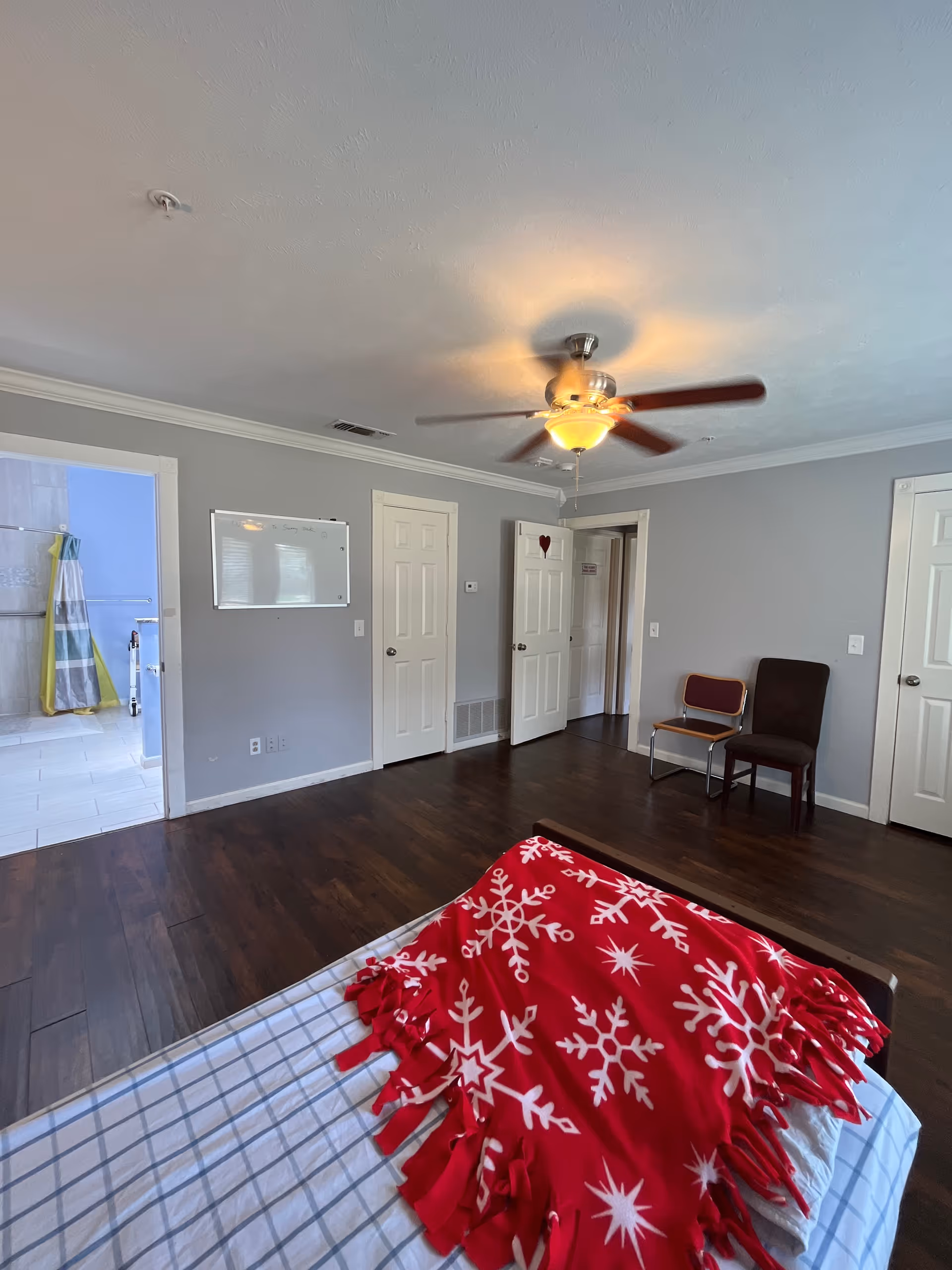 A bedroom with a bed, ceiling fan, two chairs, and an open bathroom doorway.