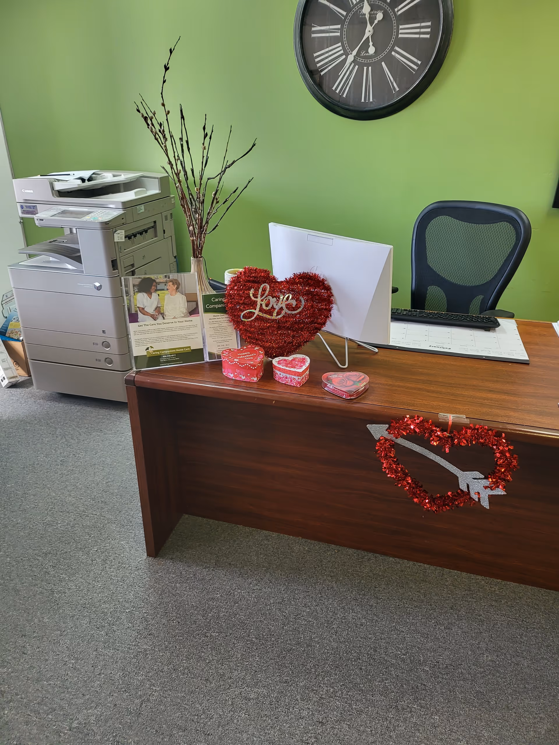 Reception desk with computer, printer, heart decorations, and a green wall clock in lobby