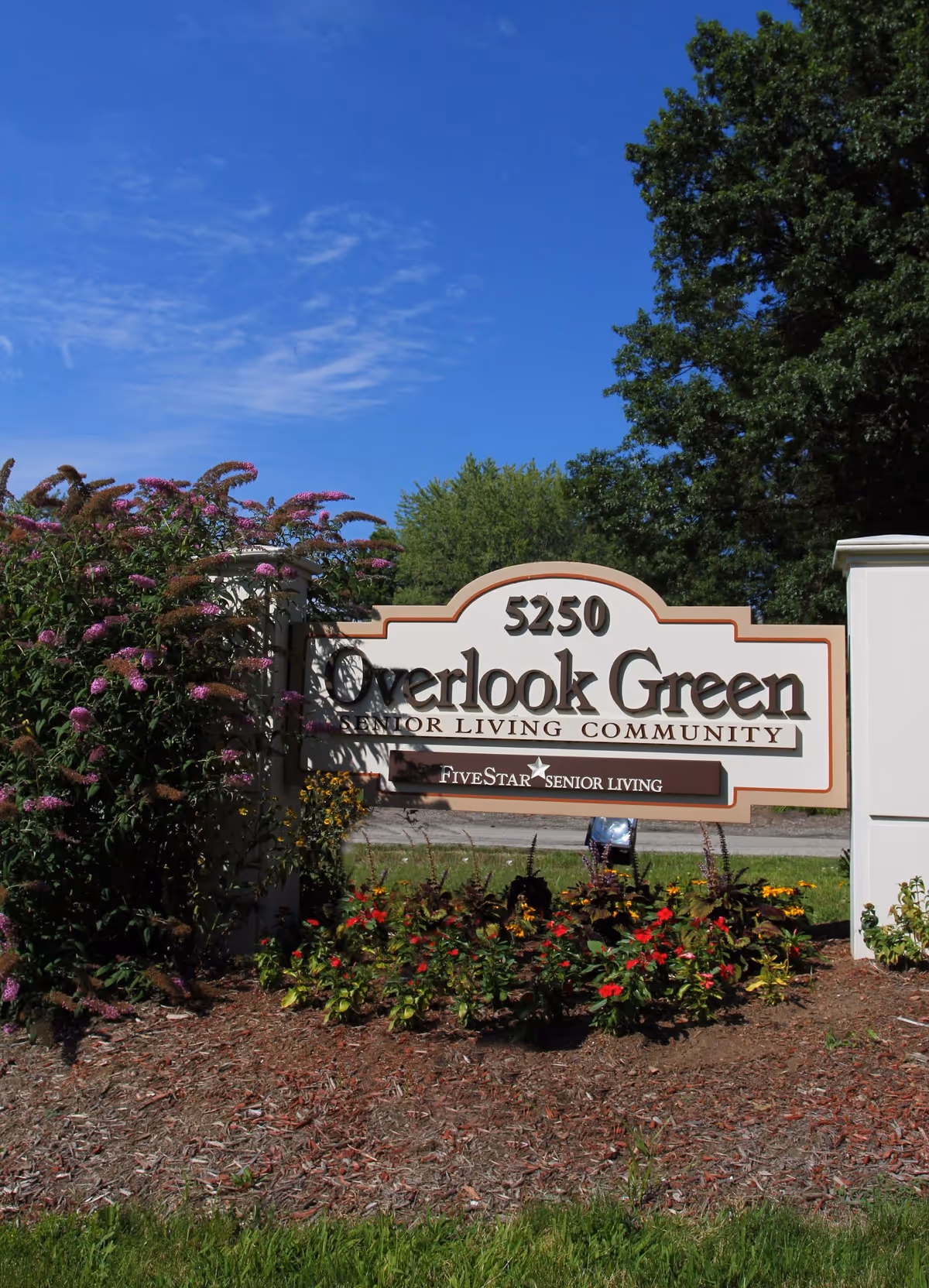 Exterior sign reading Overlook Green at entrance, surrounded by flowers and trees under a blue sky
