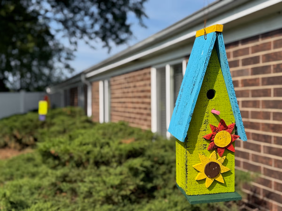 Colorful birdhouse decor hangs outside a brick facility with shrubs nearby.