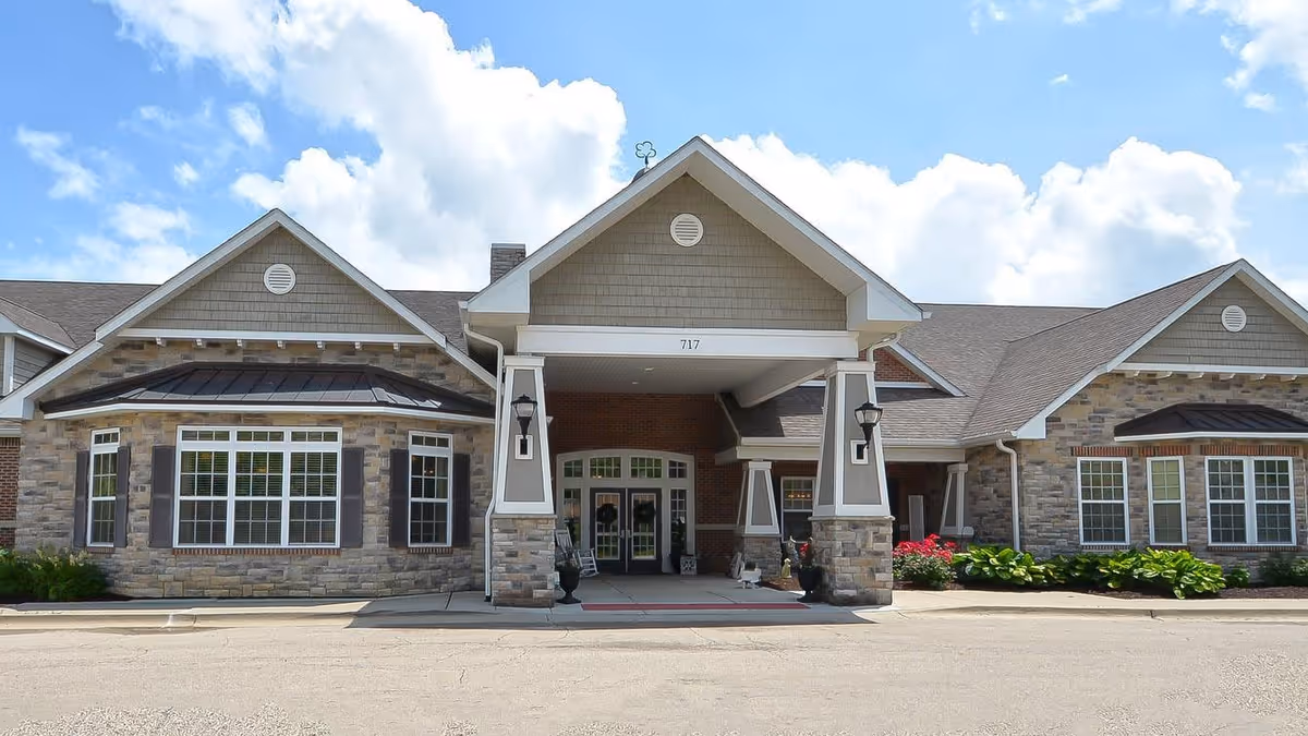 Front entrance of a single-story senior living facility with a covered porte-cochere, stone and brick facade, and landscaped beds under a blue sky.