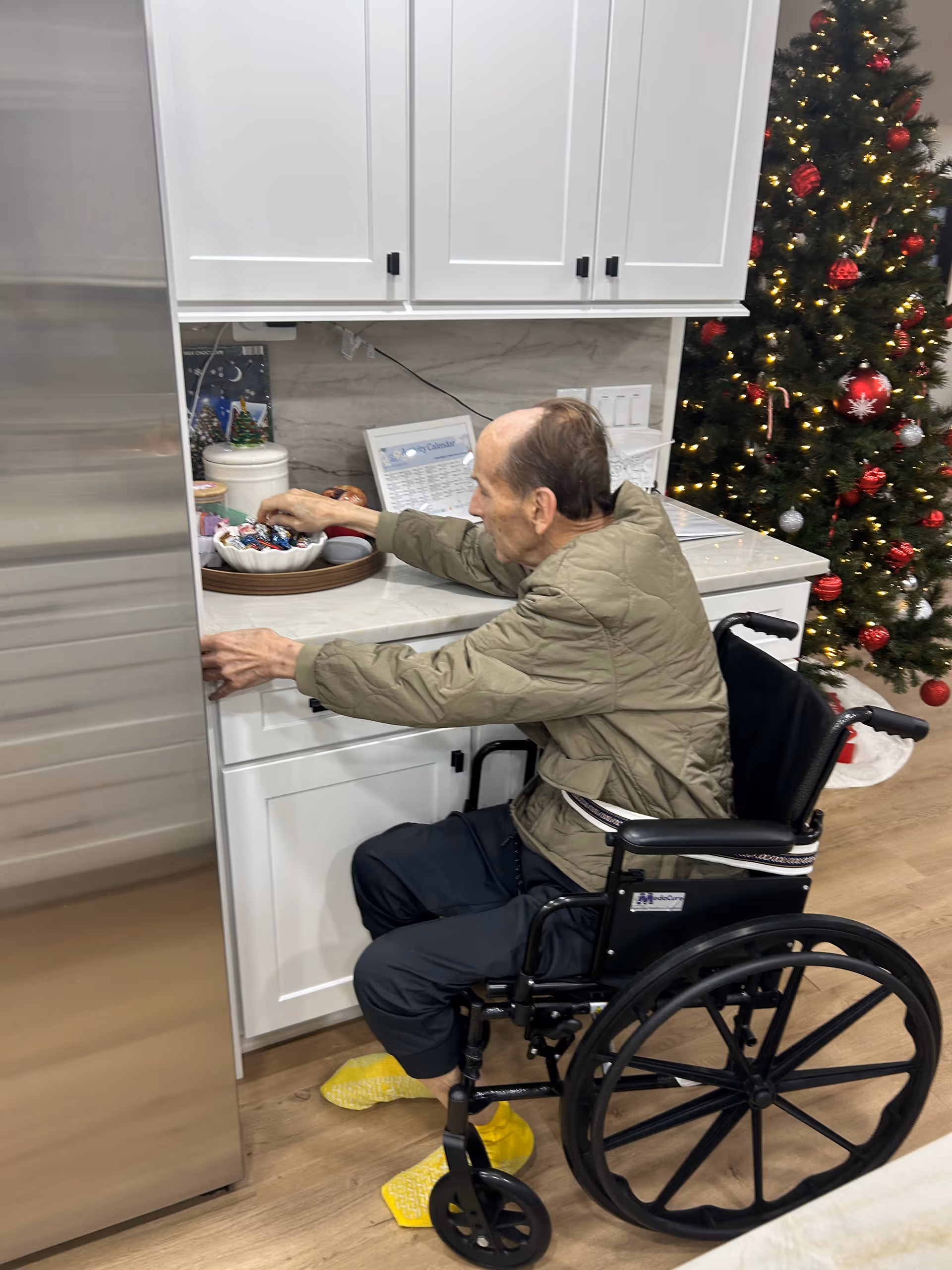 Elderly man in a wheelchair reaches into white kitchen cabinets near a Christmas tree