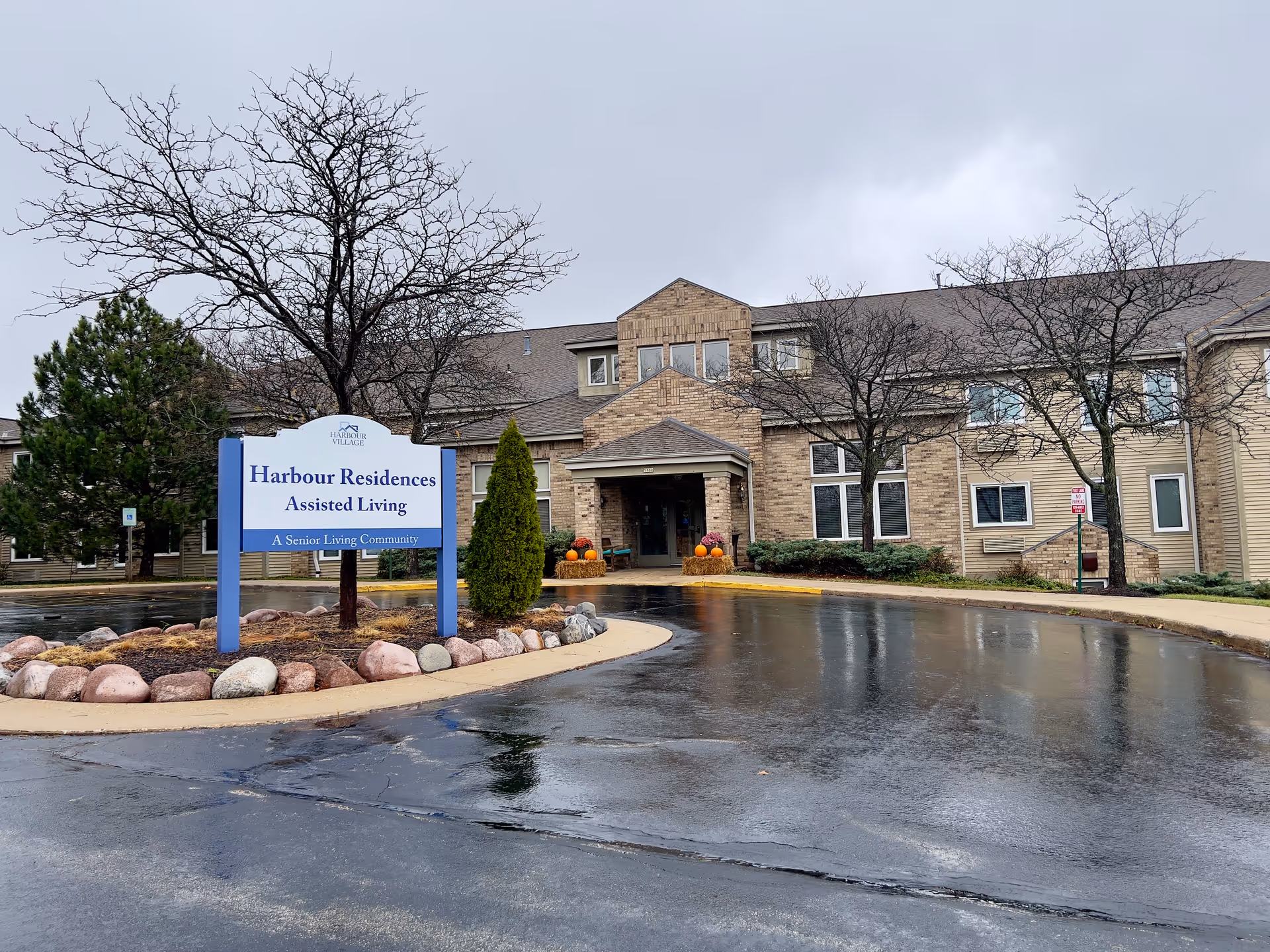 Front entrance of Harbour Residences assisted living building with its sign, wet driveway, and seasonal decorations on an overcast day.