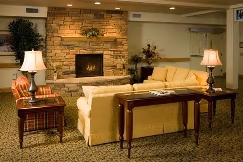 A cozy living room area with a stone fireplace in the center, flanked by a beige sofa and a plaid armchair. Two wooden side tables with lamps are placed beside the seating, and a wooden console table is behind the sofa. The room has warm lighting and a patterned carpet.