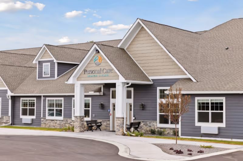 Front exterior of a single-story gray senior living building with a covered entry, benches, and a curved driveway.