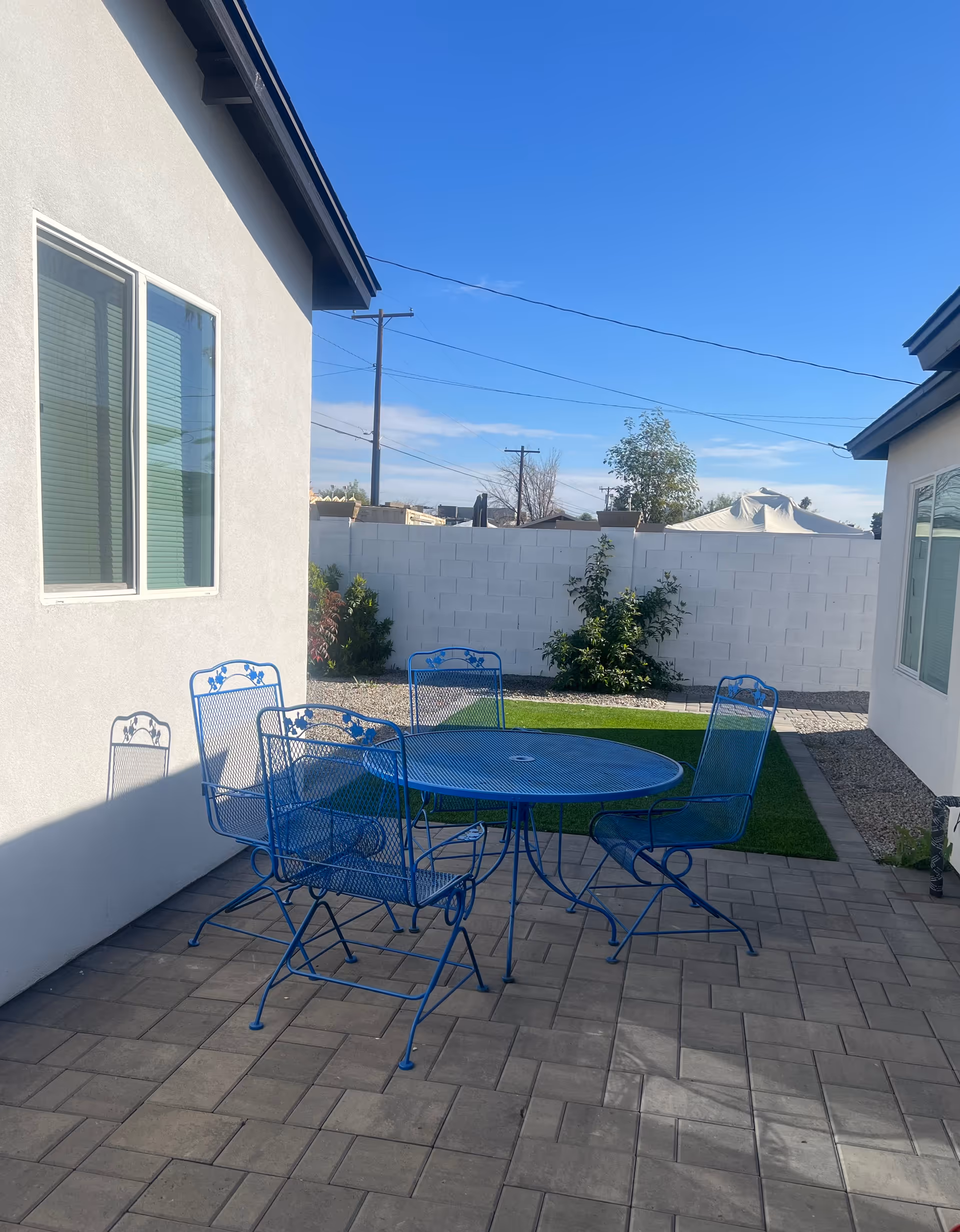 Outdoor patio area with a blue metal table and four matching chairs on a tiled floor, surrounded by white walls and a small patch of green grass with some plants and trees in the background under a clear blue sky.