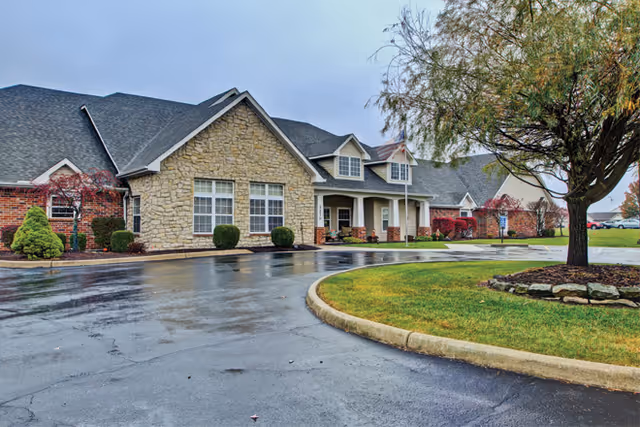 Stone and brick Brookdale Buck Creek entrance, curved driveway, and manicured trees outside.