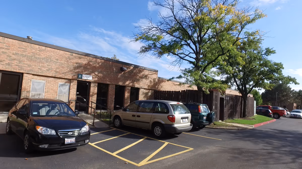 Brick senior living facility exterior with parked cars and a tree-lined lot.