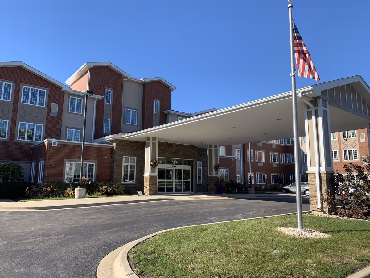 Front entrance of a multi-story brick senior living building with a covered porte-cochere and an American flag.