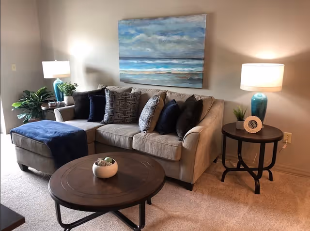 A cozy living room area with a beige sectional sofa adorned with multiple dark and patterned throw pillows. There is a round wooden coffee table in front of the sofa with a small decorative bowl on it. On either side of the sofa are round wooden side tables, each with a blue ceramic lamp and small plants. A large painting of a seascape hangs on the wall above the sofa. The room has beige carpeting and neutral-colored walls.