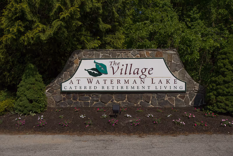 Stone sign at the entrance of The Village at Waterman Lake, a catered retirement living facility, surrounded by greenery and small flowers planted in front.