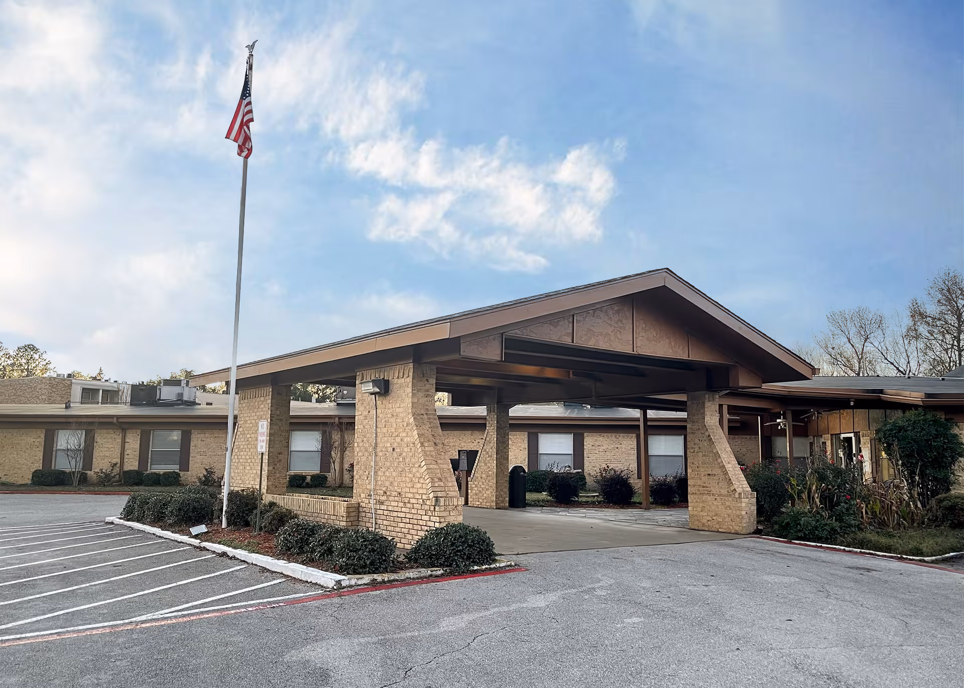 Front entrance of a single-story brick senior living facility with a covered porte-cochere, an American flag on a flagpole, and a parking area.