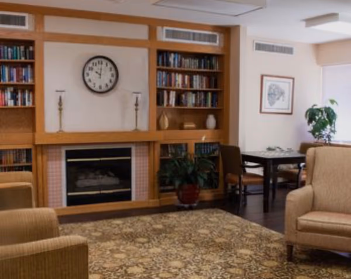 Cozy lounge area with bookshelves, fireplace, clock, plants, and seating.